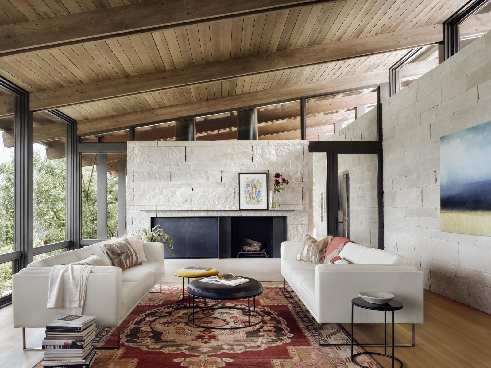 Interior living room, comprised of stone wall and fireplace, wooden ceiling and beams, floor to ceiling windows and clerestory windows The linear plans of the main house are comprised of a simple stone “box” of rooms that anchor the structures to the hillside, living room is connected to the landscape.