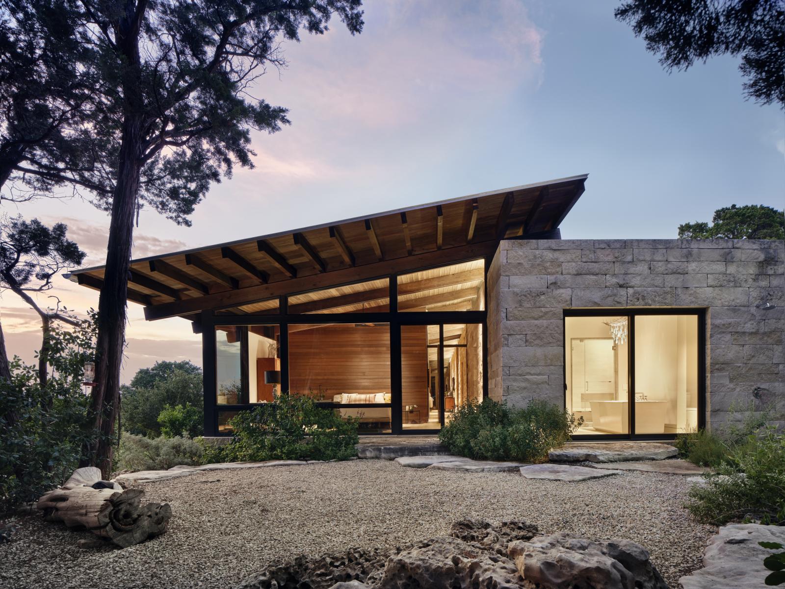 Exterior evening dusk shot, sun is setting and interior is glowing Rainwater collection tanks and an aerobic treatment system are used for irrigation. The roof photovoltaic system generates electricity. A ground source heat pump provides the heat and A/C